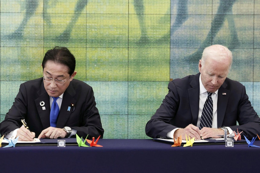 Japan's Prime Minister Fumio Kishida and President Joe Biden sit at a bench looking down as they write.