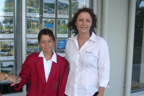 A young boy stands with his mother smiling outside real estate business. He is wearing a red jacket and she has on a white shirt