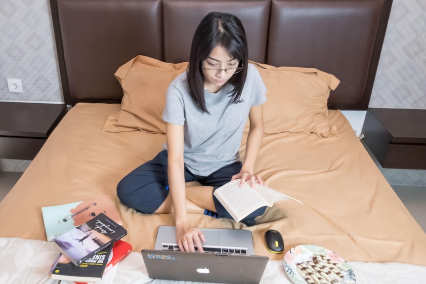 Una donna seduta sul suo letto davanti al suo laptop con libri da un lato, un piatto di cibo dall'altro e un libro aperto in grembo.
