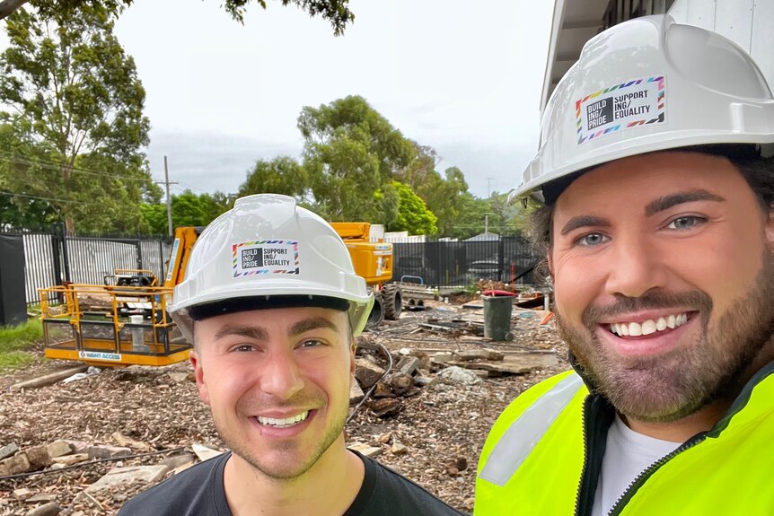 Two young men smiling and wearing safety helmets on a construction site.