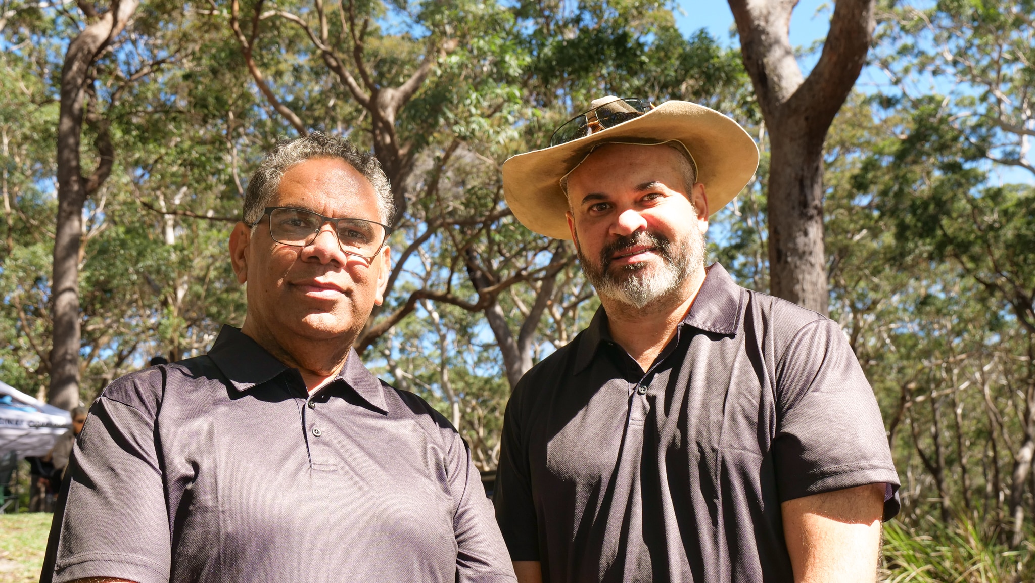 Двое мужчин стоят в окружении высоких деревьев, улыбаются, оба коренные жители, в коричневых рубашках, один в шляпе.