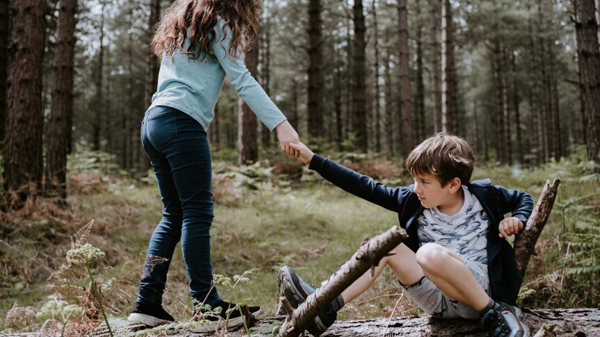 Two children help each other up in the woods.