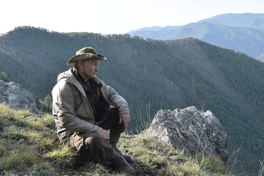 Vladimir Putin sits on the side of a hill with mountain ranges in the background.