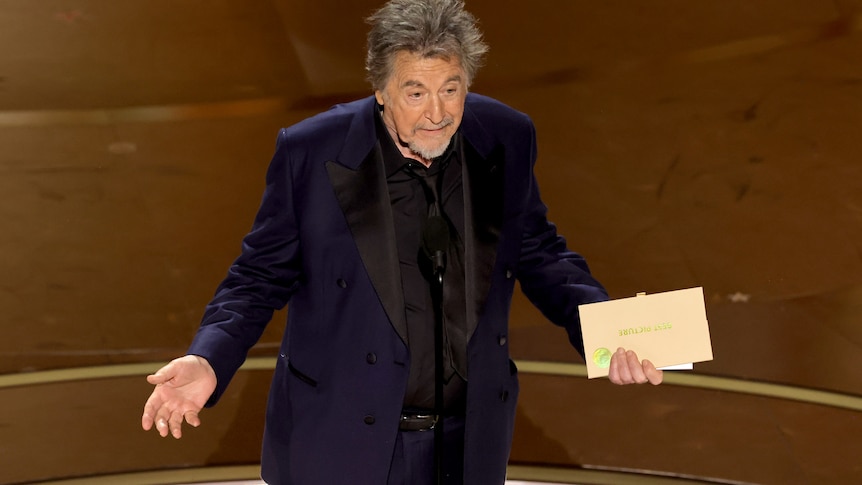 A man in a blue suit holds an envelope in his left hand.