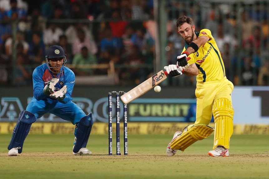 A helmetless Glenn Maxwell advances down the pitch to slap a white cricket ball while MS Dhoni watches on