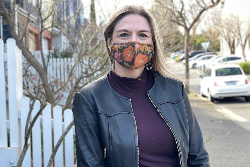 Susan stands, with mask on, in an inner-city suburban street in winter daylight.