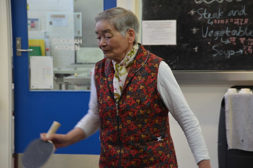 A lady with a paddle for table tennis in her hand, a blackboard in the background offering food.