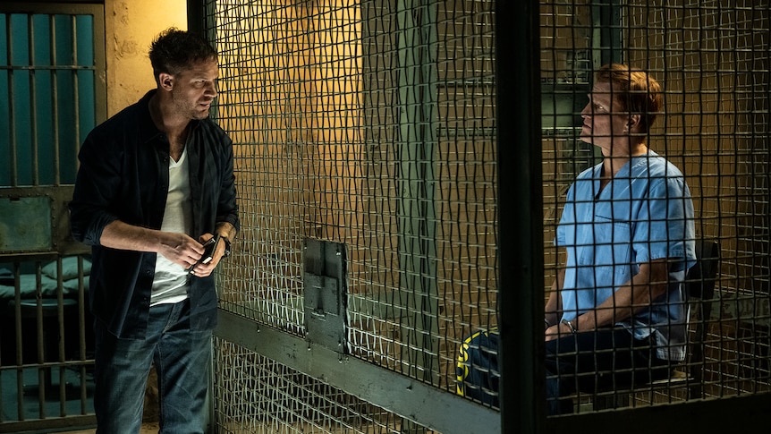 Man with scruffy dark hair and stubble wears white shirt and dark jacket outside prison cell in which a blonde man sits cuffed.