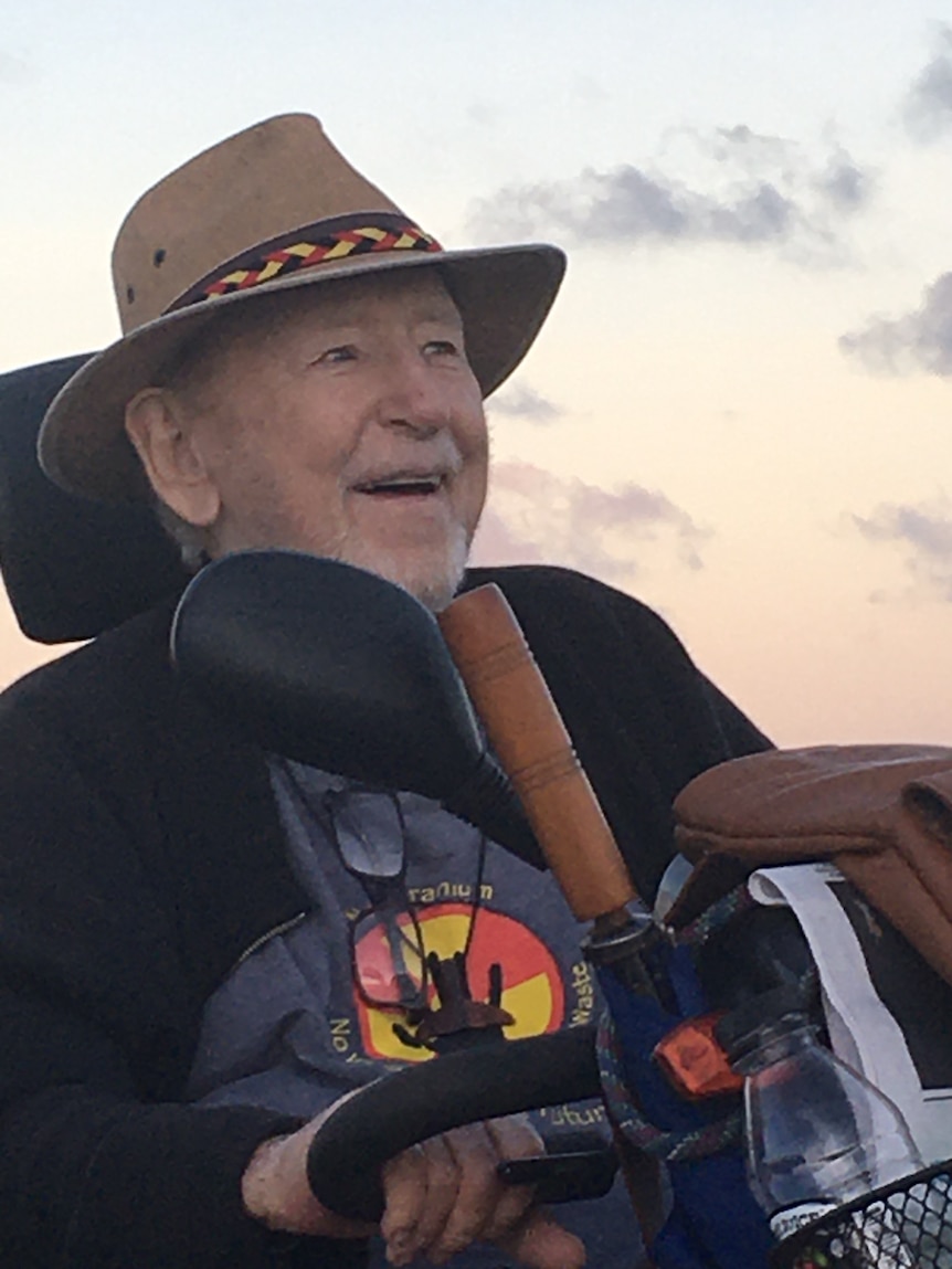 A man in a wheelchair wearing a hat smiles with the sun setting behind him