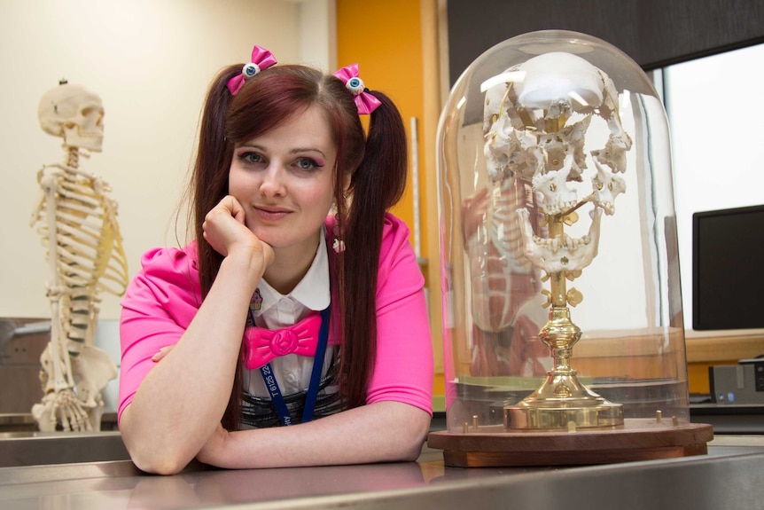 Hannah Lewis leans on a workbench in the lab, next to a model of a skull inside a glass dome.
