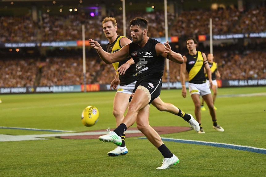 An AFL player kicks the football as a grandstand packed with fans can be seen in the distance.