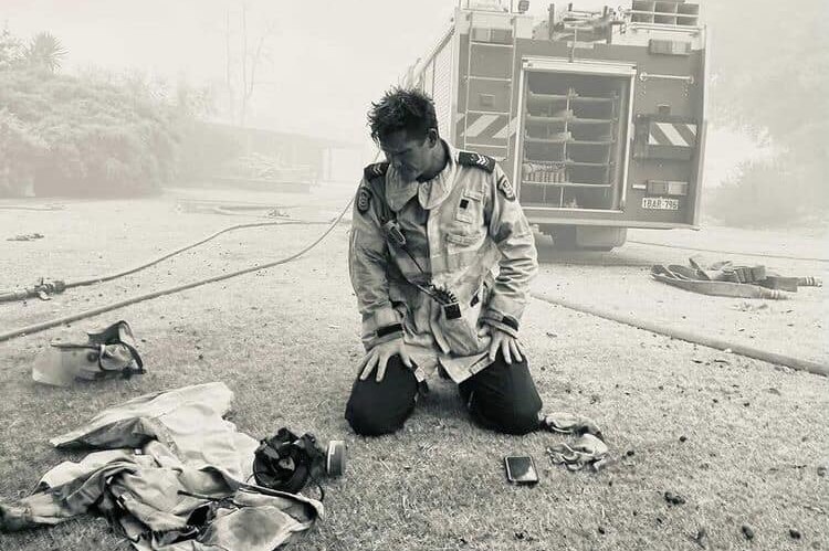 A firefighter kneels at the back of a fire truck, a respirator in front of him.