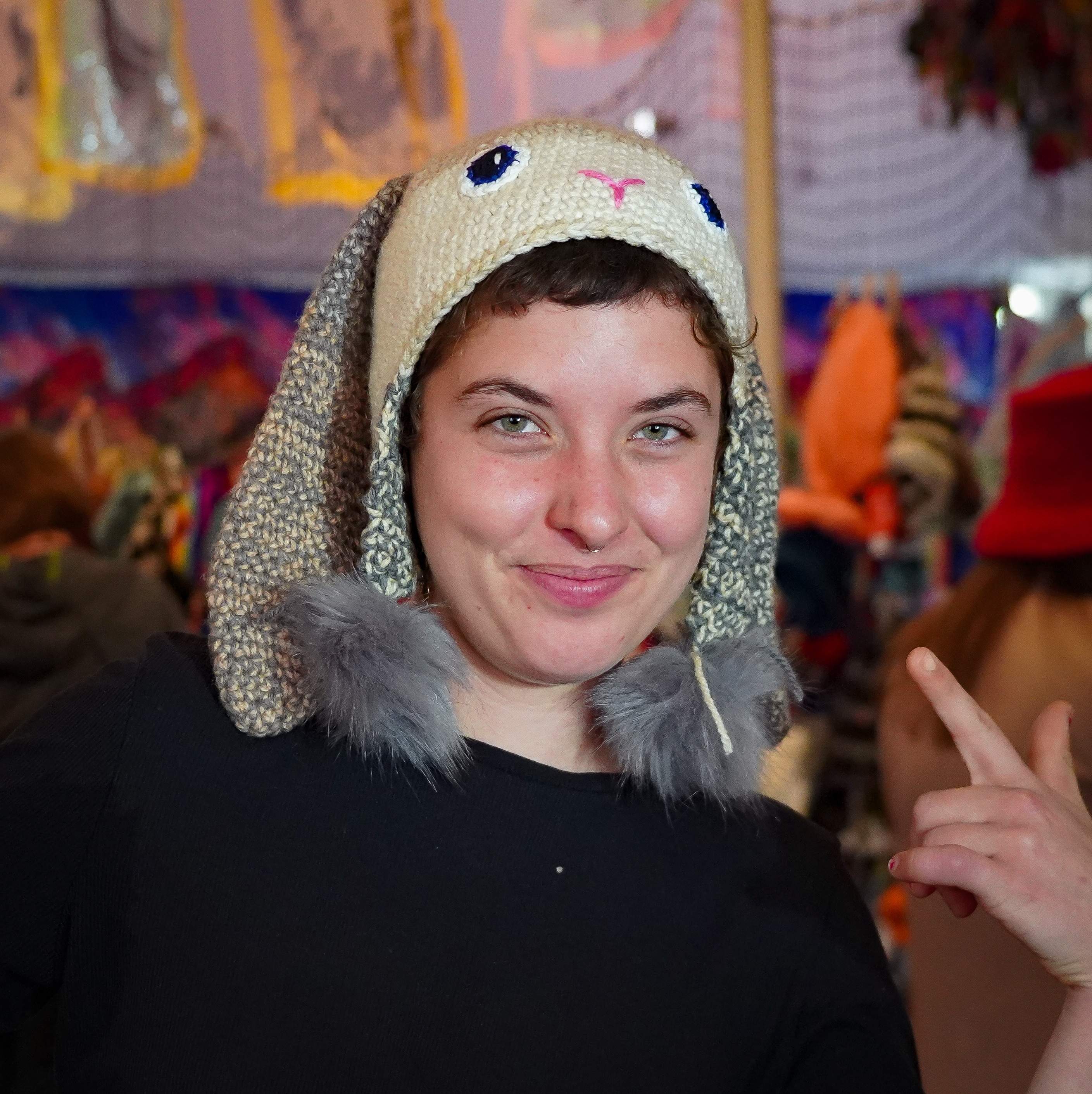 A woman wears a white and grey knitted beanie, there's blue eyes and bunny ears on the top of the hat.