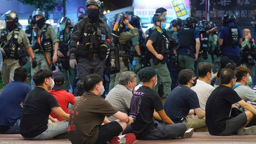 More than a dozen men in surgical masks sit in front of a large number of police in riot gear.