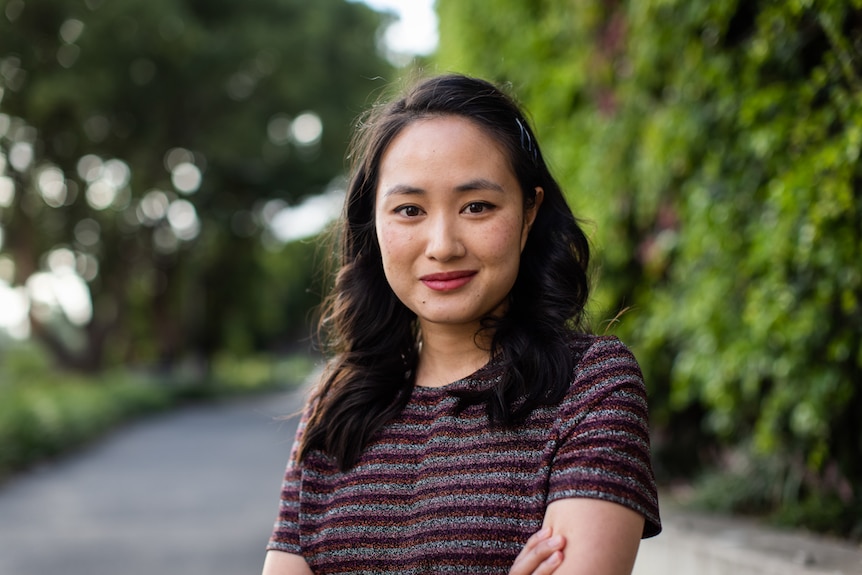 Young woman with shoulder-length wavy black hair wearing striped sparkling top looks at camera smiling, with hedge in background