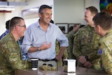 A middle-aged man in a shirt standing at a table talking with three soldiers who are seated