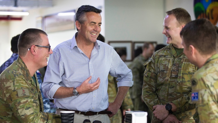 A middle-aged man in a shirt standing at a table talking with three soldiers who are seated