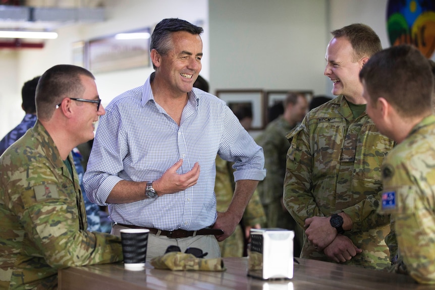 A middle-aged man in a shirt standing at a table talking with three soldiers who are seated