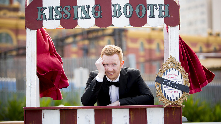 Comedian Luke McGregor waits awkwardly at at kissing booth in the ABC TV series Luke Warm Sex.