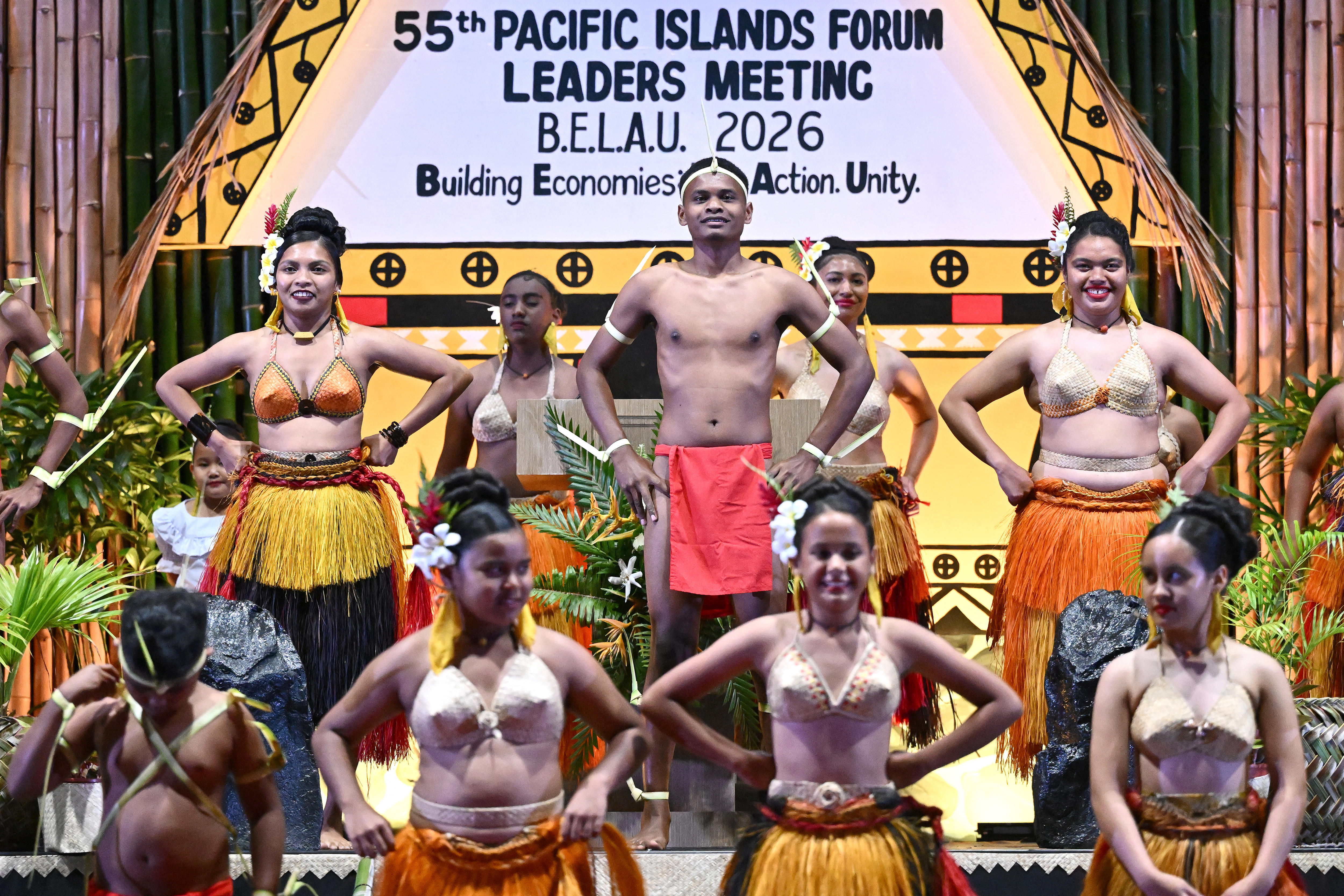 Nine men and women, wearing traditional outfits, stand in rows on a stage.