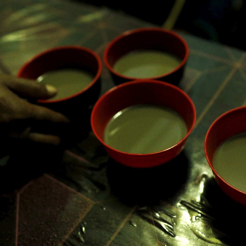 Close-up of a hand reaching out to four bowls of brown liquid kava on a table.