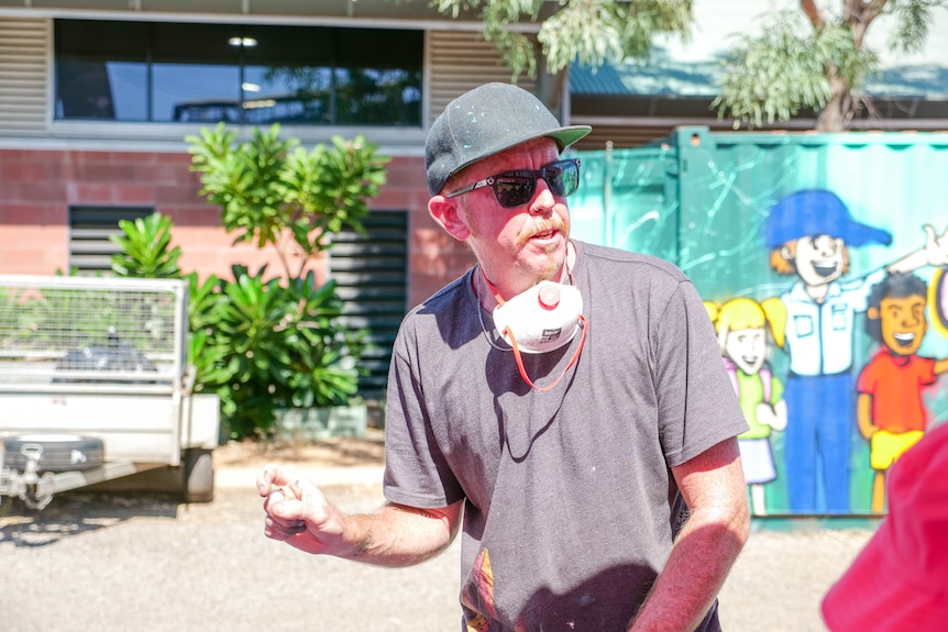 A man with a mask around his chin wearing a cap and sunglasses talks.