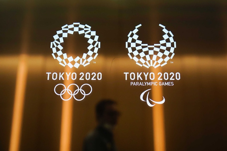 A silhouetted man walking past logos for the Tokyo Olympics and Paralympics, with light reflected.