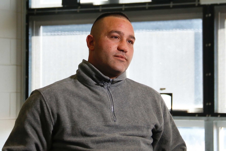 A man in a grey jumper with windows behind him.