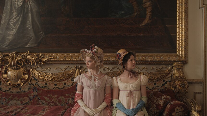 Two women in Regency England era costumes sit on large ornate red sofa with gold-leaf detailing, in front of large oil painting.
