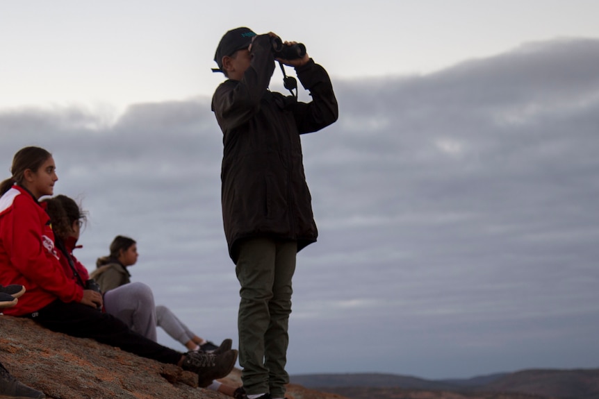 four kids sit on a boulder, one stands. All are holding or using binoculars and looking off camera.