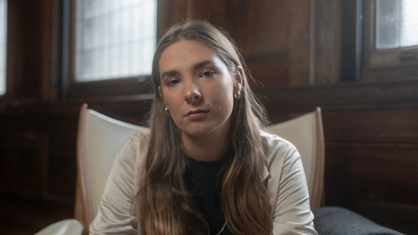 Evelyn Araluen sits in a white chair, leaning slightly forward, with her hair tucked behind her ears, looking into the camera