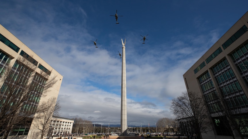 Four helicopters fly in formation over a tall statue with a fork at the top of it in between two large buildings