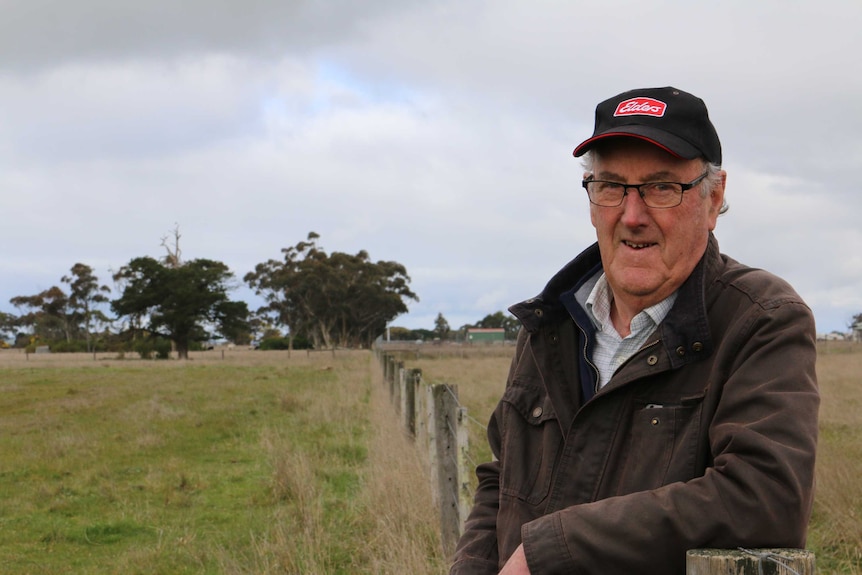 A man wearing a hat leaning against a fence post in a paddock.