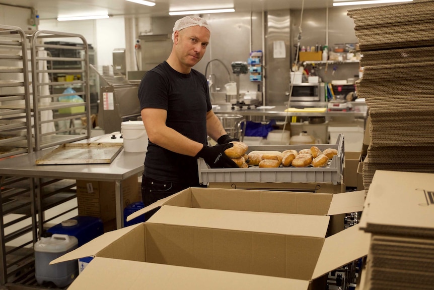 A man in a black t-shirt and gloves and wearing a hair net is packing bread into a box from inside a commercial kitchen.