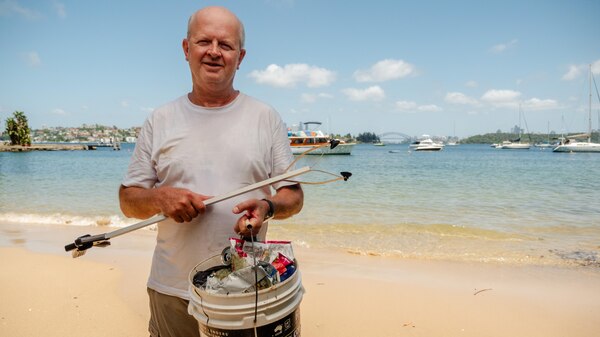 A middle-aged man stands on the beach holding a bucket and rubbish tongs. 