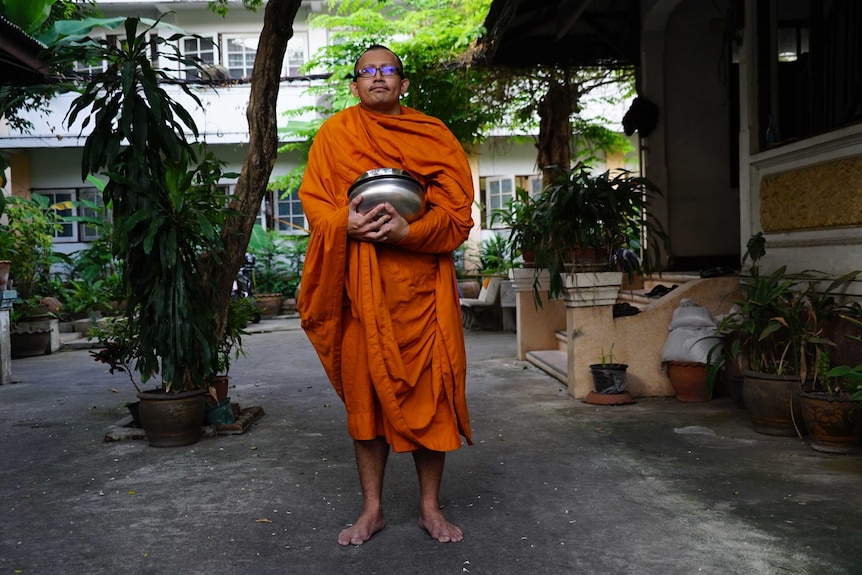 An Asian man with short hair and glasses and dressed in a saffron robe clutches a large metal bowl with both hands