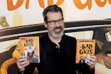 Middle-aged white man with short grey hair, beard and glasses wears black suit and stands on red carpet in front of movie poster