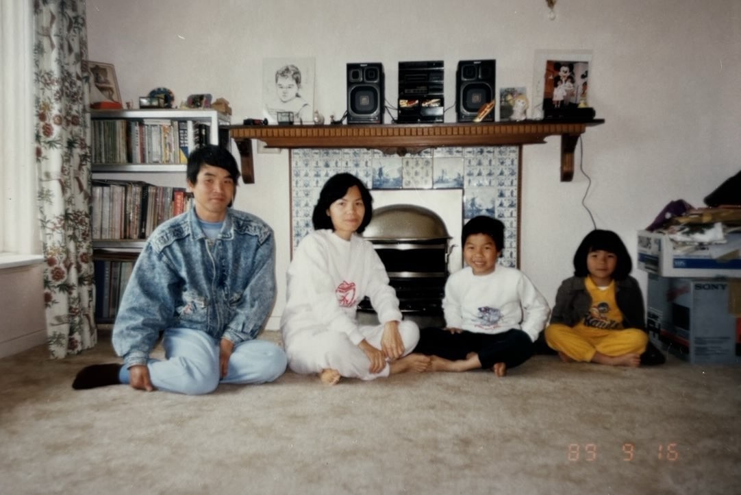 Uma imagem retrô de uma família de quatro pessoas, dois pais, um menino e uma menina sentados em um tapete em frente à lareira, no chão acarpetado, sorrindo.