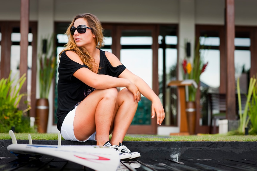 Tyler Wright sitting on the ground next to her surfboard with sunglasses on