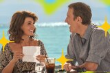 Toula from My Big Fat Greek Wedding sits at a table with her husband smiling at him, the ocean behind them