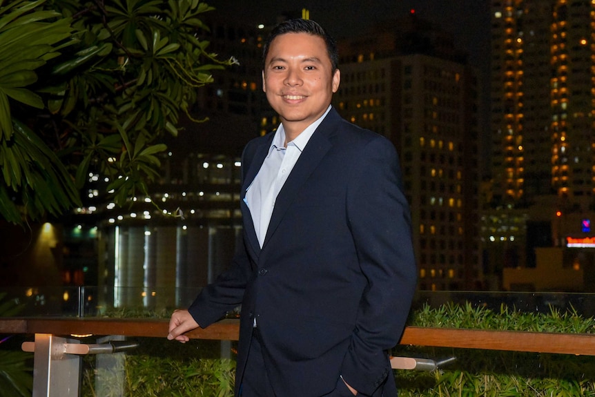 A man wearing a suit stands smiling at the camera at night-time with city lights behind him.