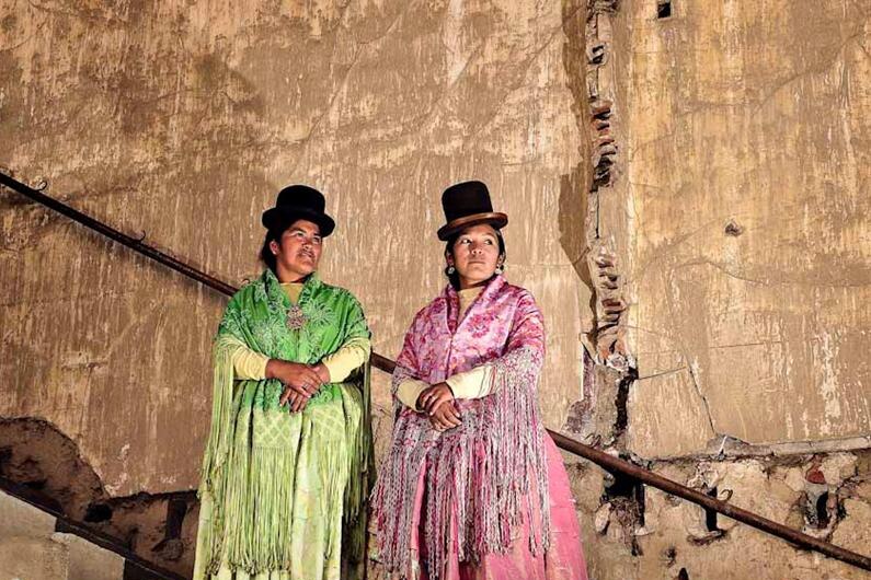 Two Flying Cholitas wearing colourful costumes pose on a set of worn-looking steps.