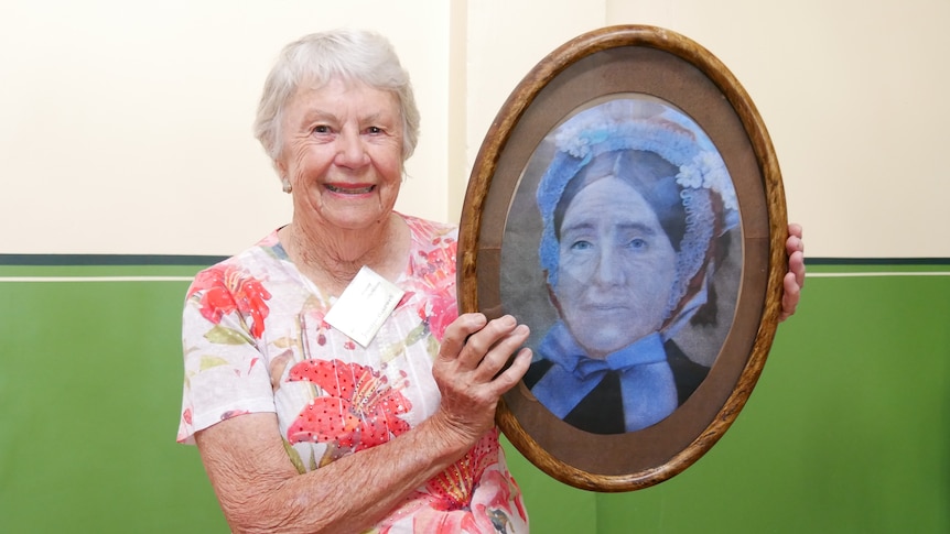 An older woman holds a framed photo of a convict woman.