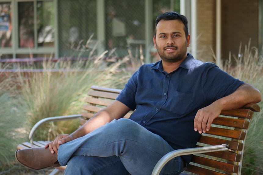 a man in a blue shirt and jeans sits on a park bench portrait