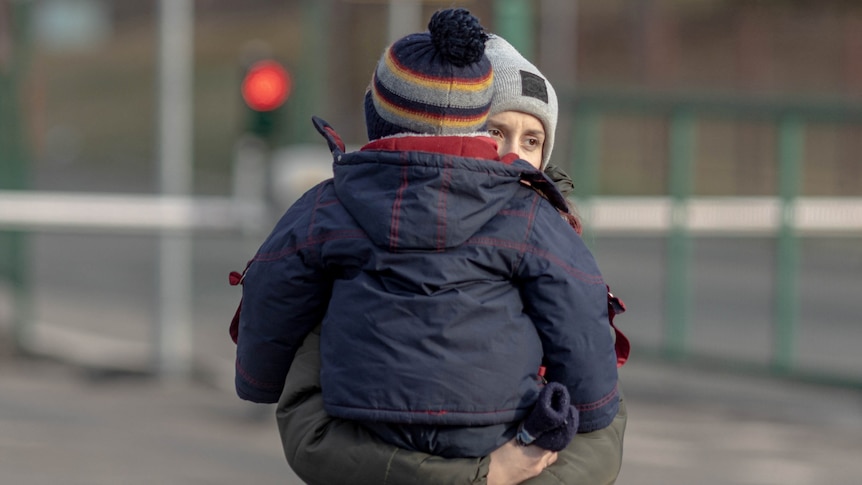 A woman wearing a beanie holds a small child in her arms walking along a street.