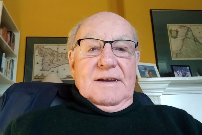 An older man with a bald head and glasses sitting in a room with gold coloured walls.