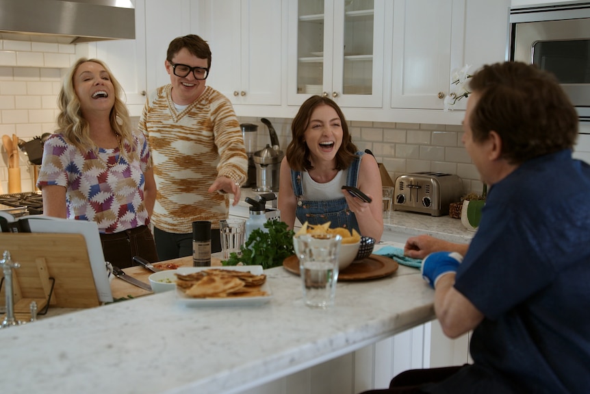 A couple and their two adult children gather around a kitchen bench in a large house, laughing together.