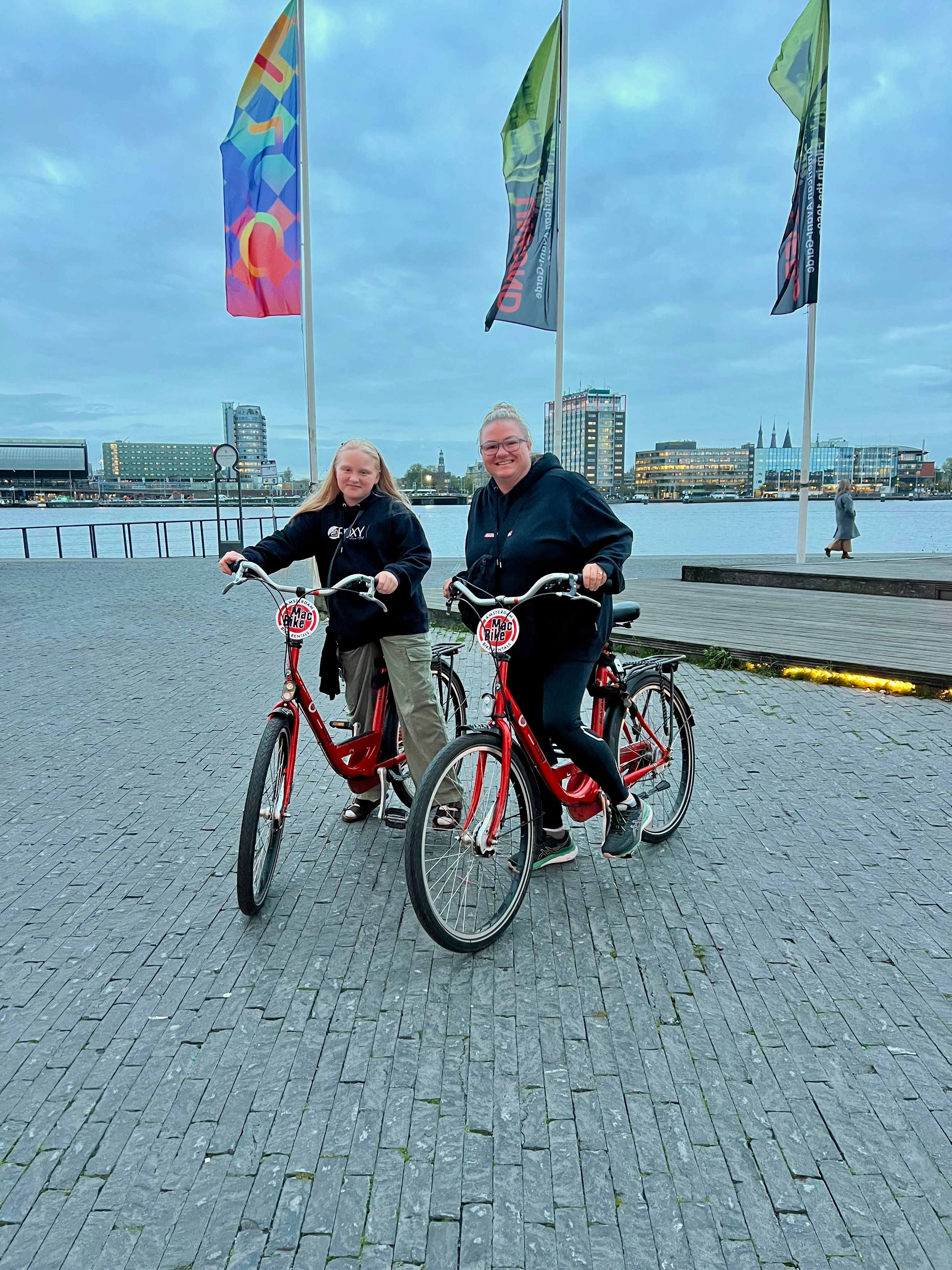 A woman and a young girl on bicycles on a paved riverside space, colourful flags fluttering behind them. 