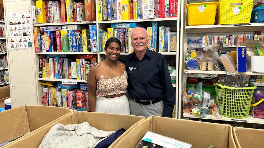 A photograph of two smiling volunteers in front of a book shelf full of books and games