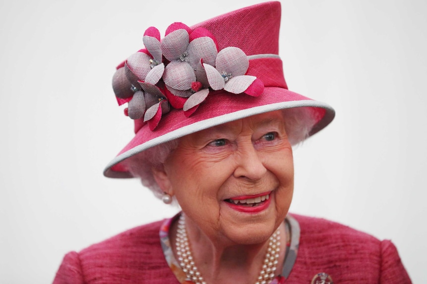 Queen Elizabeth II, wearing a pink floral hat, smiles for a photo.